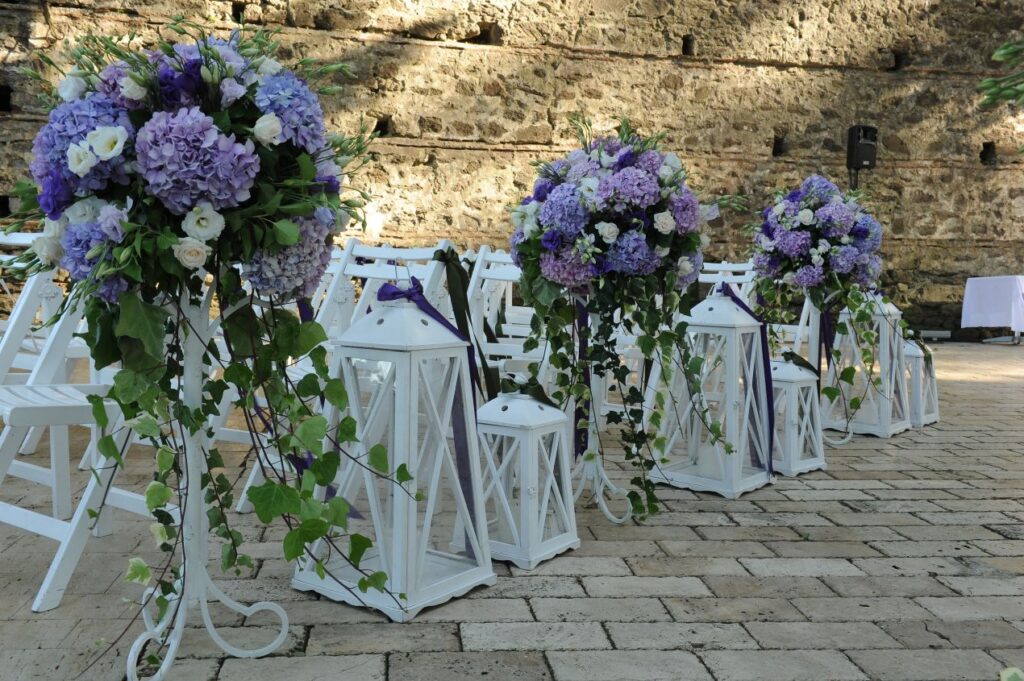 Weisse Holzstuehle mit Blumenschmuck und Laternen bei einer stilvollen Outdoor-Hochzeit – ideal fuer alle, die Stuehle mieten moechten.
